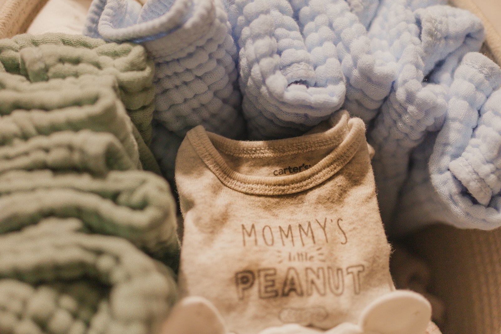 Soft folded muslin baby blankets and a onesie tucked into a woven basket, in sage and cream tones.